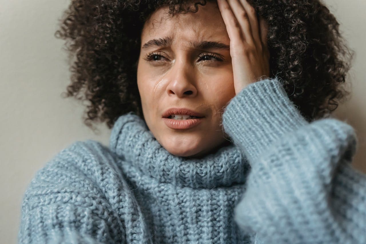 A person in tears wearing a cozy, twisted-knit blue sweater, with curly hair partially obscured by their hand against a neutral background.