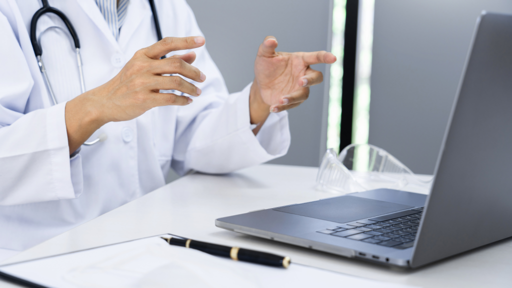 A doctor in a white coat gesturing during a video consultation on a laptop