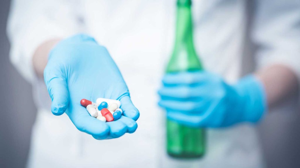 Gloved hand holding pills with a beer bottle in the background.